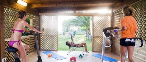 Un espacio exterior techado para gimnasio con equipo de ejercicio como cintas de correr y elípticas.
