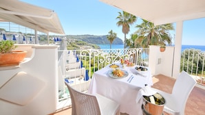 A balcony with a table set for two, overlooking a coastal landscape with palm trees and a view of the sea.