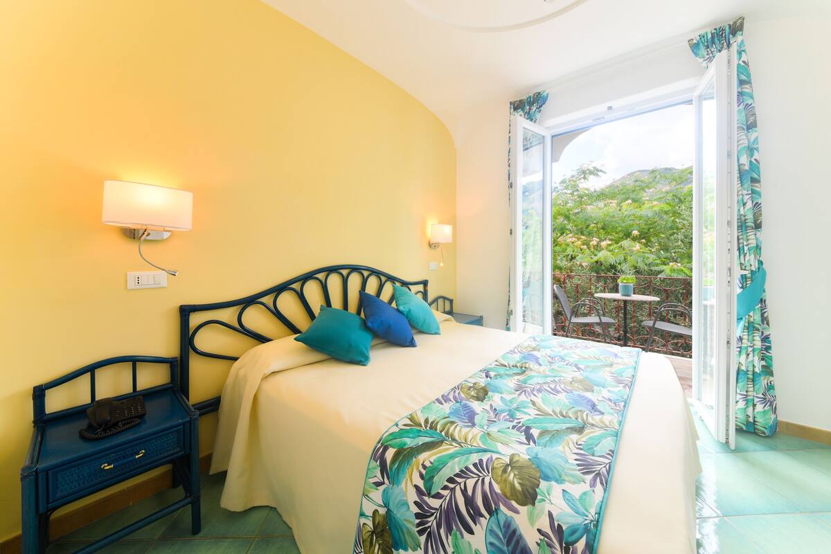 A bedroom with a bed, bedside tables, and a view of a garden through an open door.