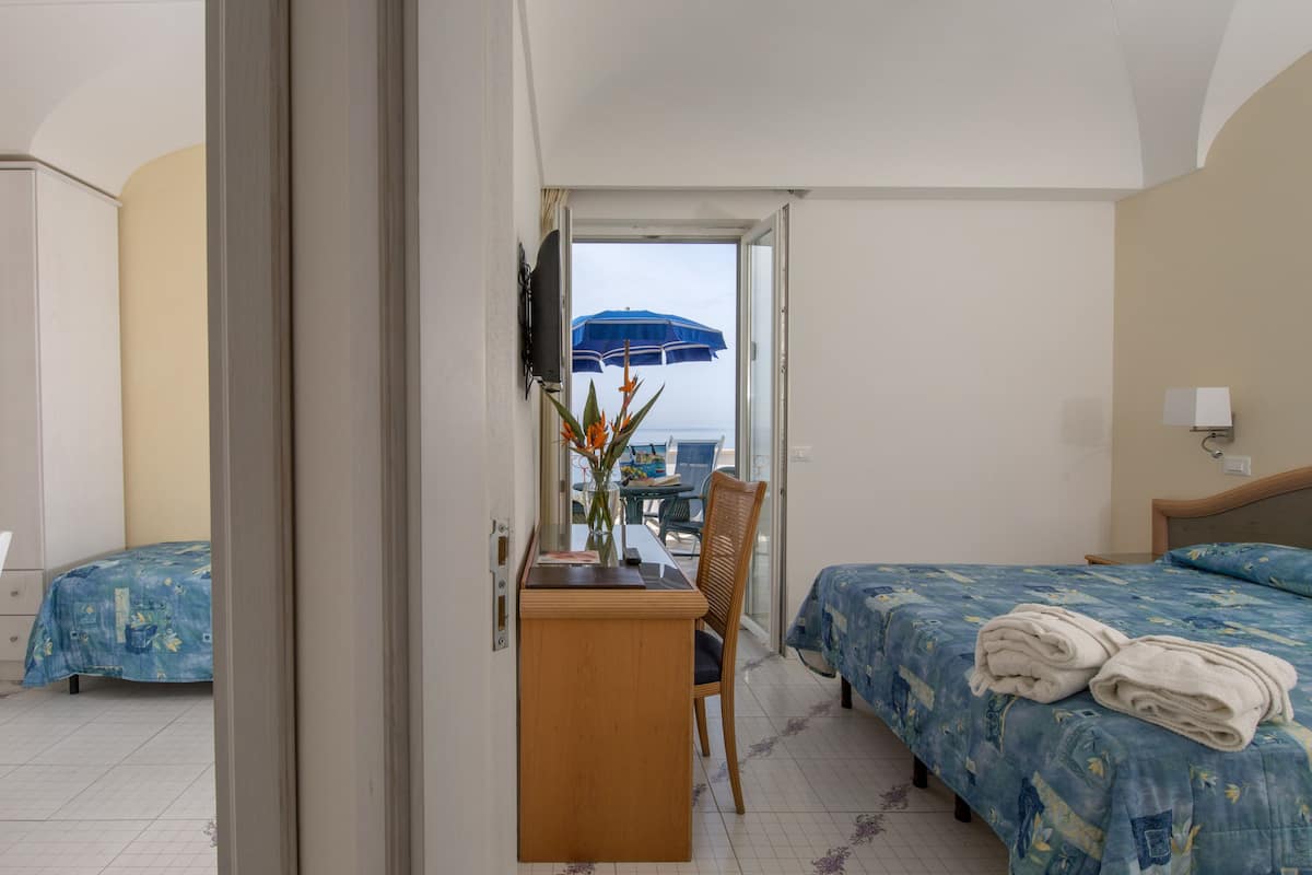 A hotel room with two beds, a desk with a chair, a TV, and a view of the sea through a window.