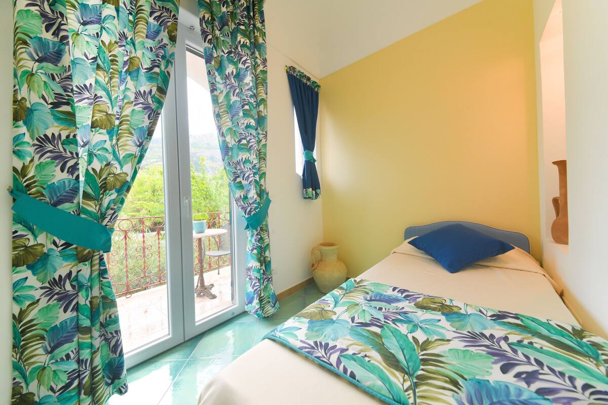 A bedroom with a bed, a window with curtains, a vase, and a view of greenery outside.