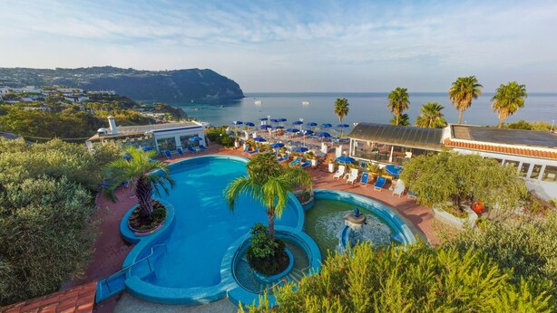 A resort with a large swimming pool, sun loungers, and a view of the sea.