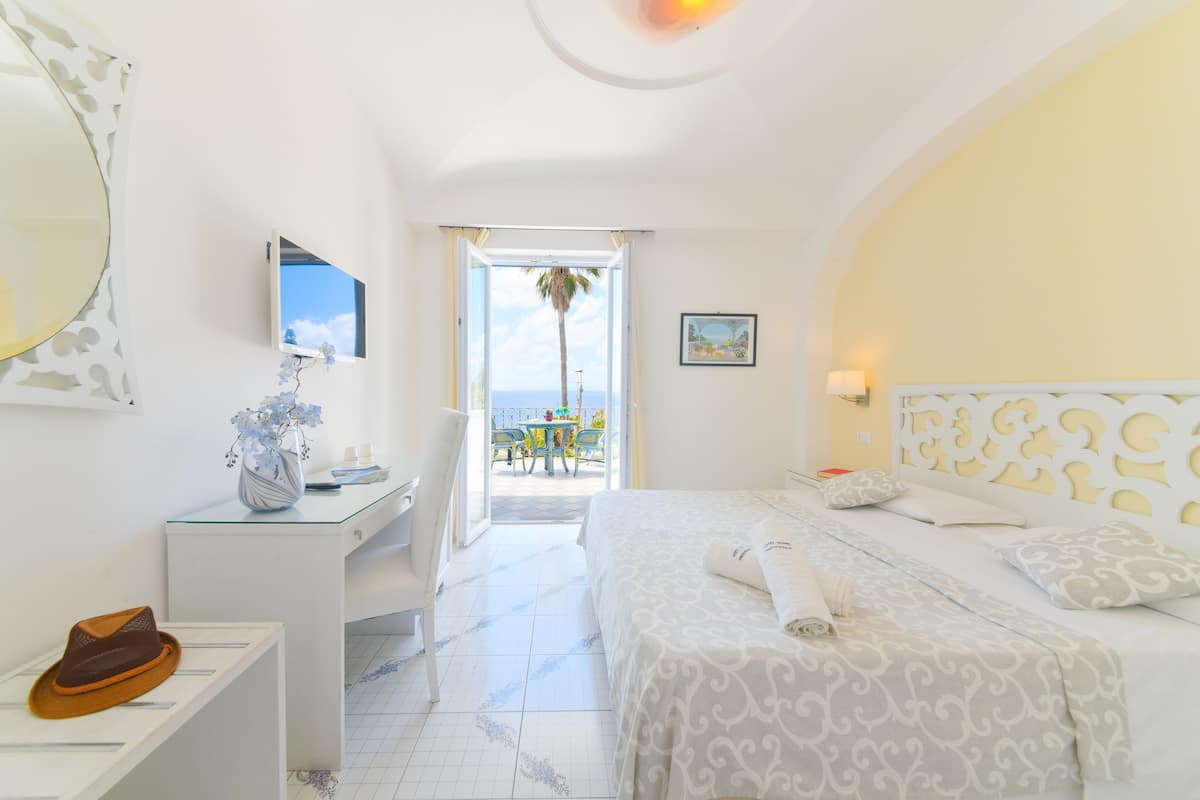 A hotel room with a bed, a desk, a chair, a vase with flowers, and a view of the sea.