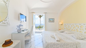 A hotel room with a bed, a desk, a chair, a vase with flowers, and a view of the sea.