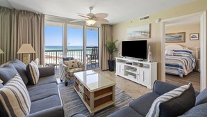 A spacious living room with a large TV, comfortable seating, a balcony with a view of the beach, and a bedroom with a bed and a painting. - Pelican Beach 0815 2 Bedroom Condo by Pelican Beach Management (Destin)