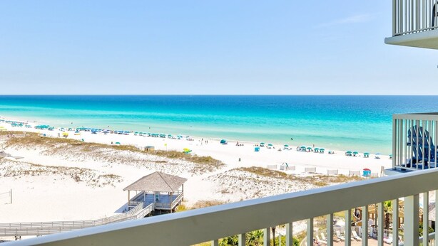 A beach view with a white railing, beach umbrellas, and a small structure on the sand. - Pelican Beach 0815 2 Bedroom Condo by Pelican Beach Management (Destin)