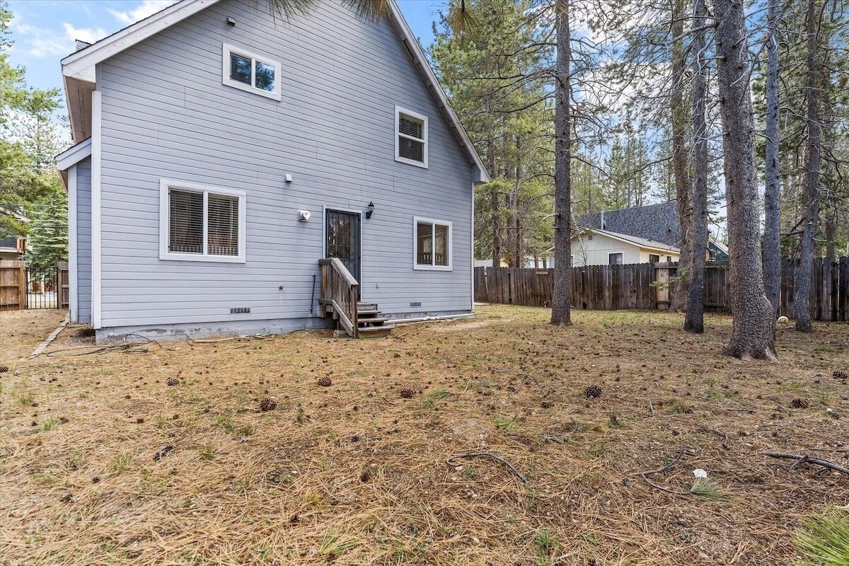 A two-story house with a front porch, a wooden fence, and a yard with pine trees and pine cones.