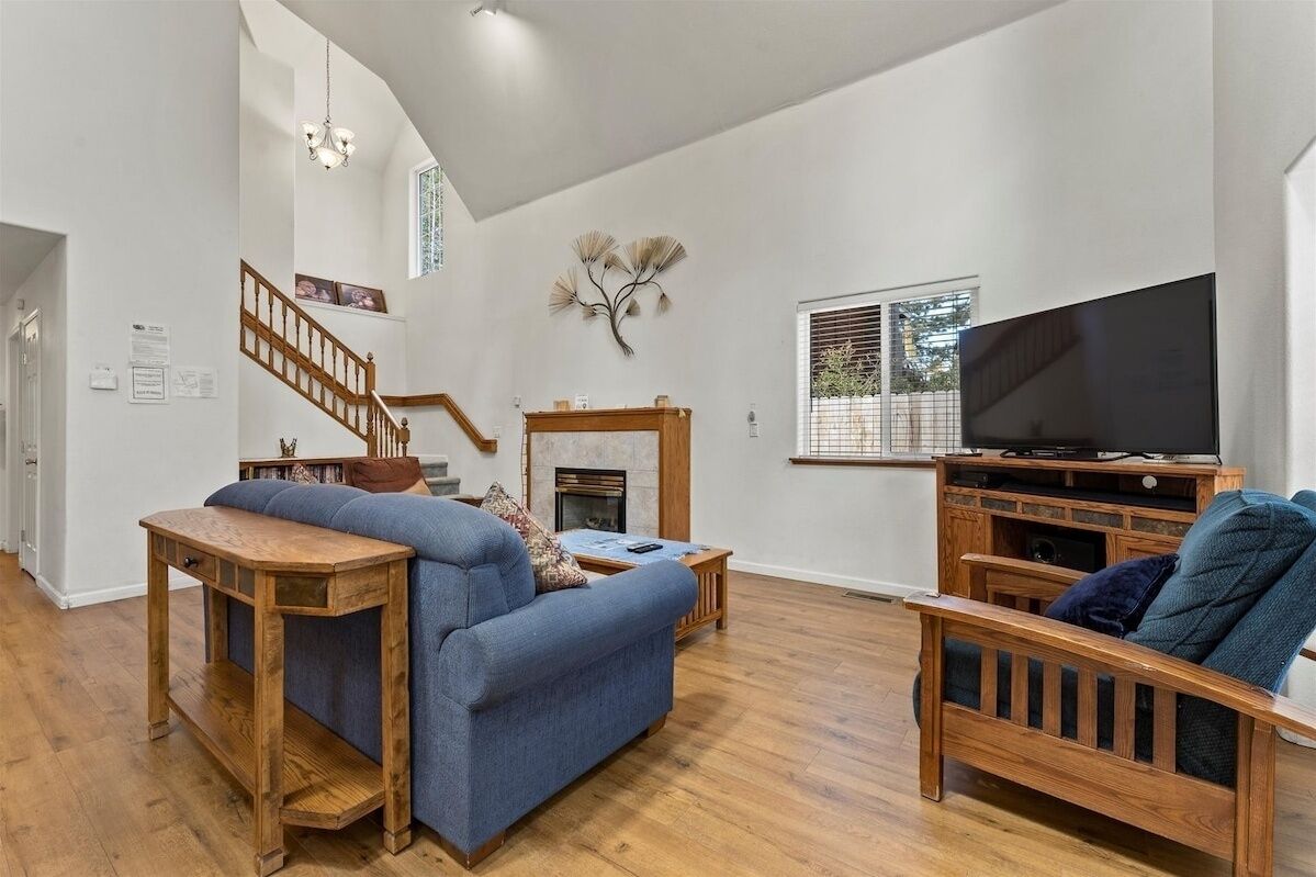A spacious living room with a blue sofa, wooden coffee table, and a TV on a wooden stand. A staircase with wooden railings leads to an upper level.