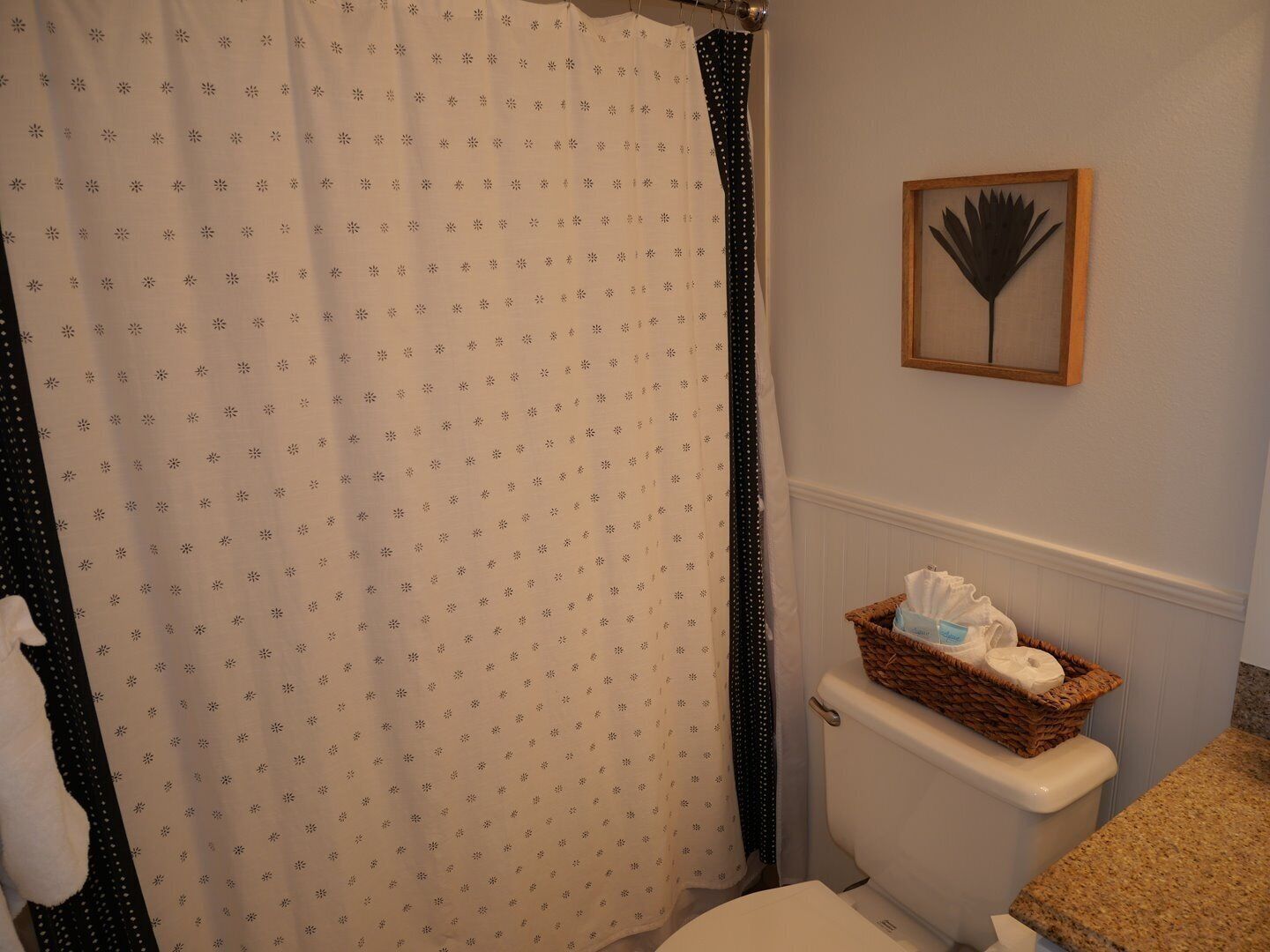 A bathroom with a shower curtain, a toilet with a basket of toiletries, and a framed artwork on the wall.