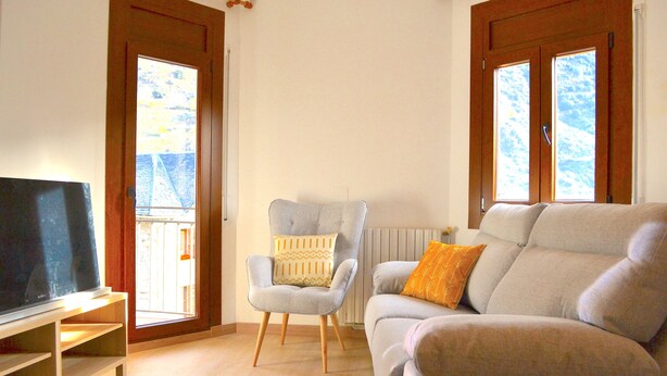 A living room with a television on a stand, a grey armchair with a patterned cushion, a beige sofa with an orange cushion, and large windows with wooden frames. - Apartment with views and central in Esterri (Esterri d'Àneu)