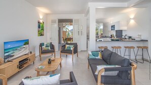 A modern living room with a TV, sofas, and a coffee table. There is a kitchen area with a bar and stools. - Beachside Starfish (Sainte-Anne)
