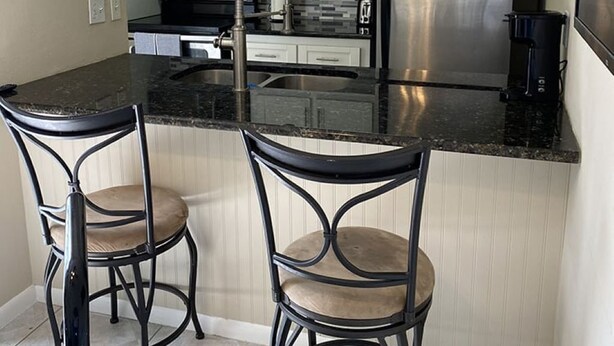 Una cocina moderna con encimera negra, dos taburetes de bar, electrodomésticos de acero inoxidable y decoración en la pared.