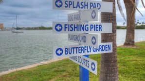 Un cartel direccional con múltiples flechas que indican diferentes lugares, como parque acuático, parque canino, muelle de pesca, parque infantil, jardín de mariposas y canchas de tenis.