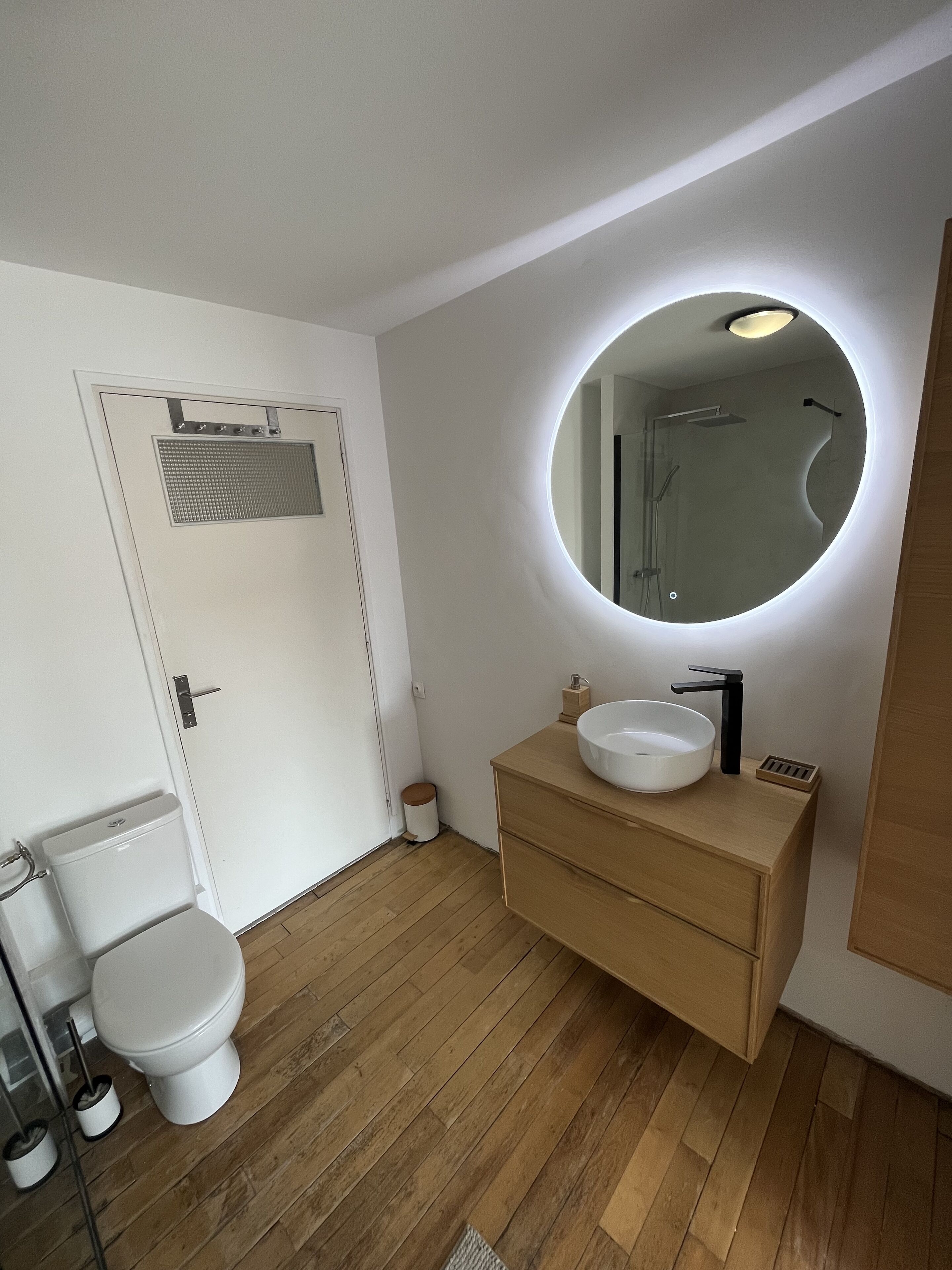 A modern bathroom with a toilet, a sink with a round mirror, and wooden flooring.