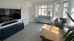 A modern living room with a flat-screen TV, a dark entertainment unit, a light-colored sofa, a wooden coffee table, and large windows offering a view of the outside. - Duplex 5 persons seaside. (Wissant)