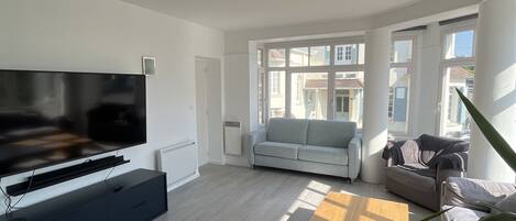 A modern living room with a flat-screen TV, a dark entertainment unit, a light-colored sofa, a wooden coffee table, and large windows offering a view of the outside.