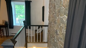 Una escalera de madera con barandillas blancas, un muro de piedra y una ventana con cortinas.