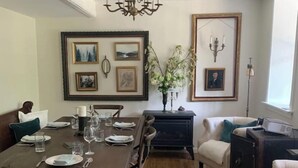 Un comedor con una mesa de madera dispuesta para cuatro personas, rodeada de sillas. Hay cuadros en las paredes, una araña y un sofá blanco.