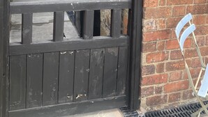 A narrow alleyway leading to a building with a stone wall and a brick section. A black gate with a metal handle is in the foreground. A sign on the brick wall reads "35 Aplington Cottage."