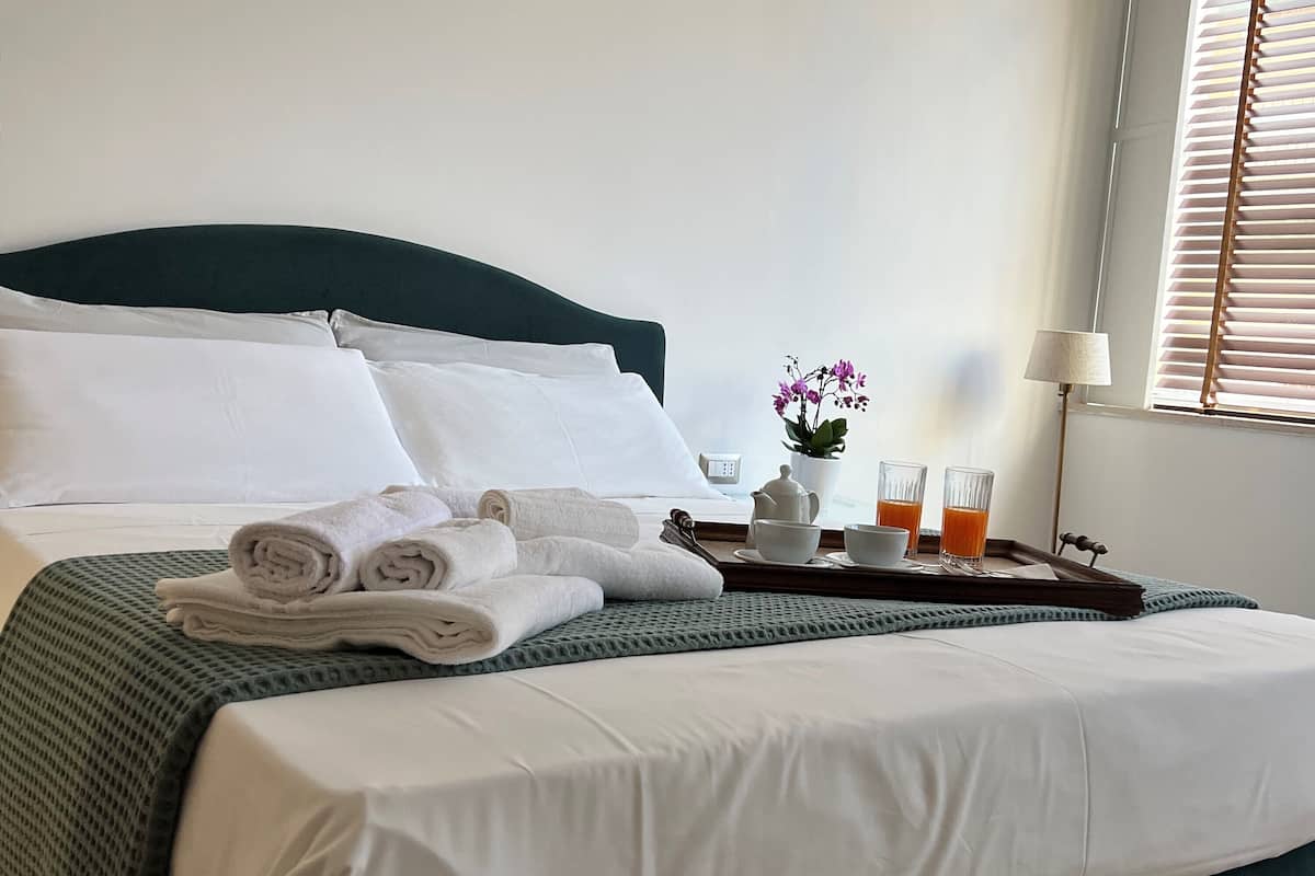Una cama bien hecha con sábanas blancas, un cabecero verde y una bandeja con toallas, un hervidor de agua y dos vasos de jugo de naranja.