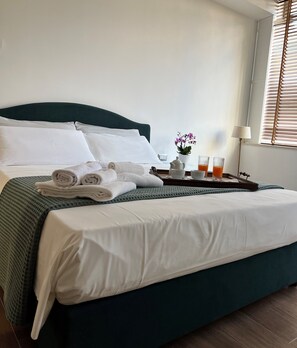 A neatly made bed with white linens, a green headboard, and a tray with towels, a teapot, and two glasses of orange juice.