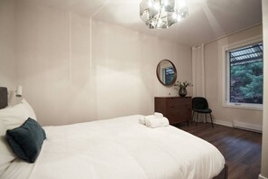 A bedroom with a bed, a dresser, a chair, a mirror, and a window with a view of greenery. - Design 3-bedroom flat with private terrace in Little Italy by Denstays (Montréal)