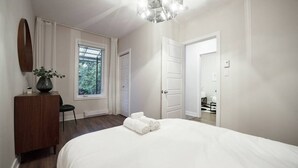 A bedroom with a large bed, a dresser, a mirror, a window with curtains, and a door leading to another room. - Design 3-bedroom flat with private terrace in Little Italy by Denstays (Montréal)