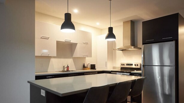 A modern kitchen with a central island, stainless steel appliances, pendant lighting, and a minimalist design. - Design 3-bedroom flat with private terrace in Little Italy by Denstays (Montréal)
