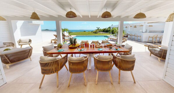 A dining area with a view of the ocean, wicker chairs, and a pool.