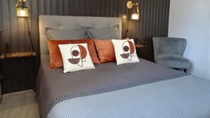 Une chambre avec un grand lit, deux lampes fixées au mur, un fauteuil gris et un panneau mural texturé.