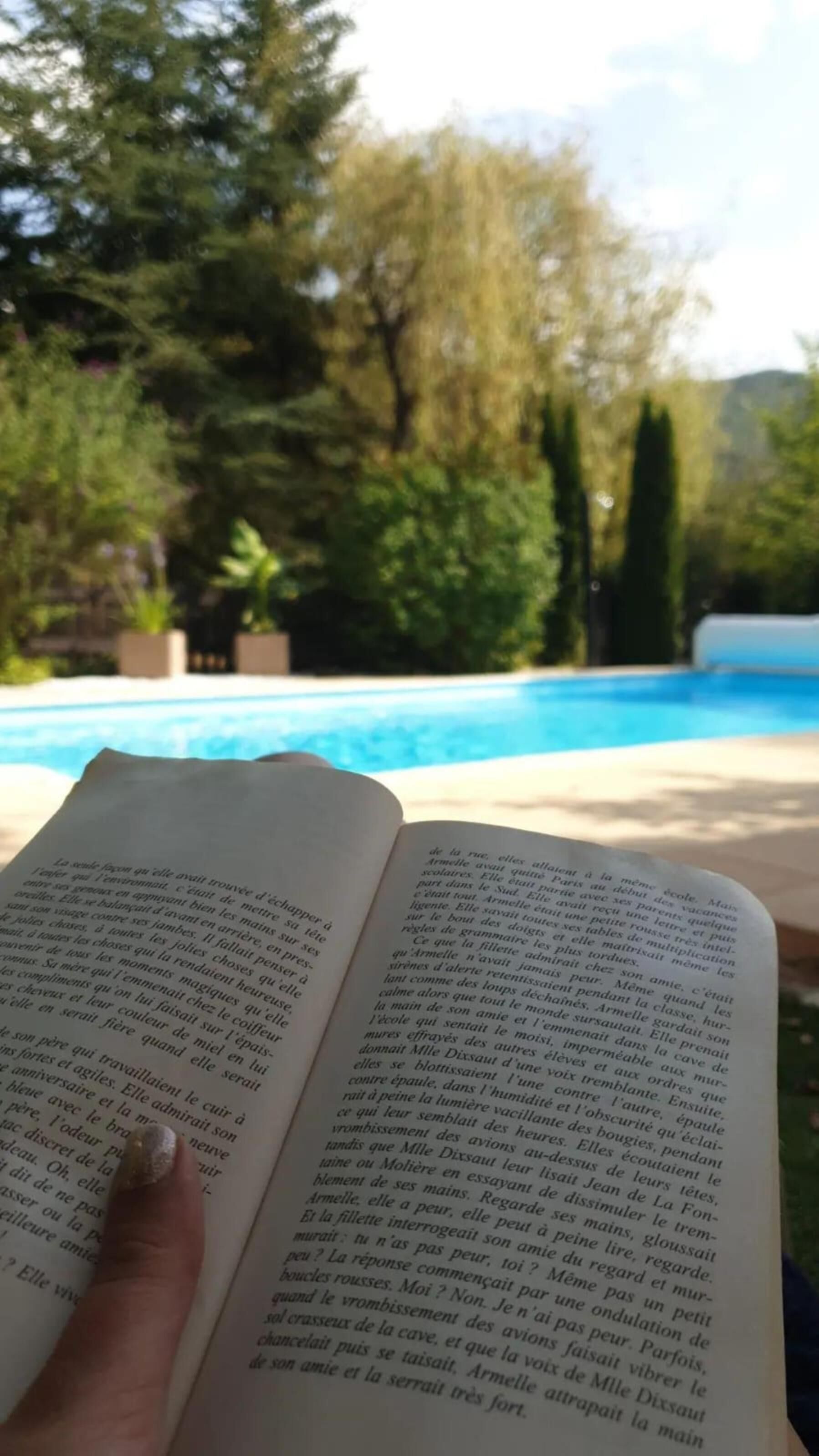 Una mano sostiene un libro abierto, con una piscina y árboles al fondo.
