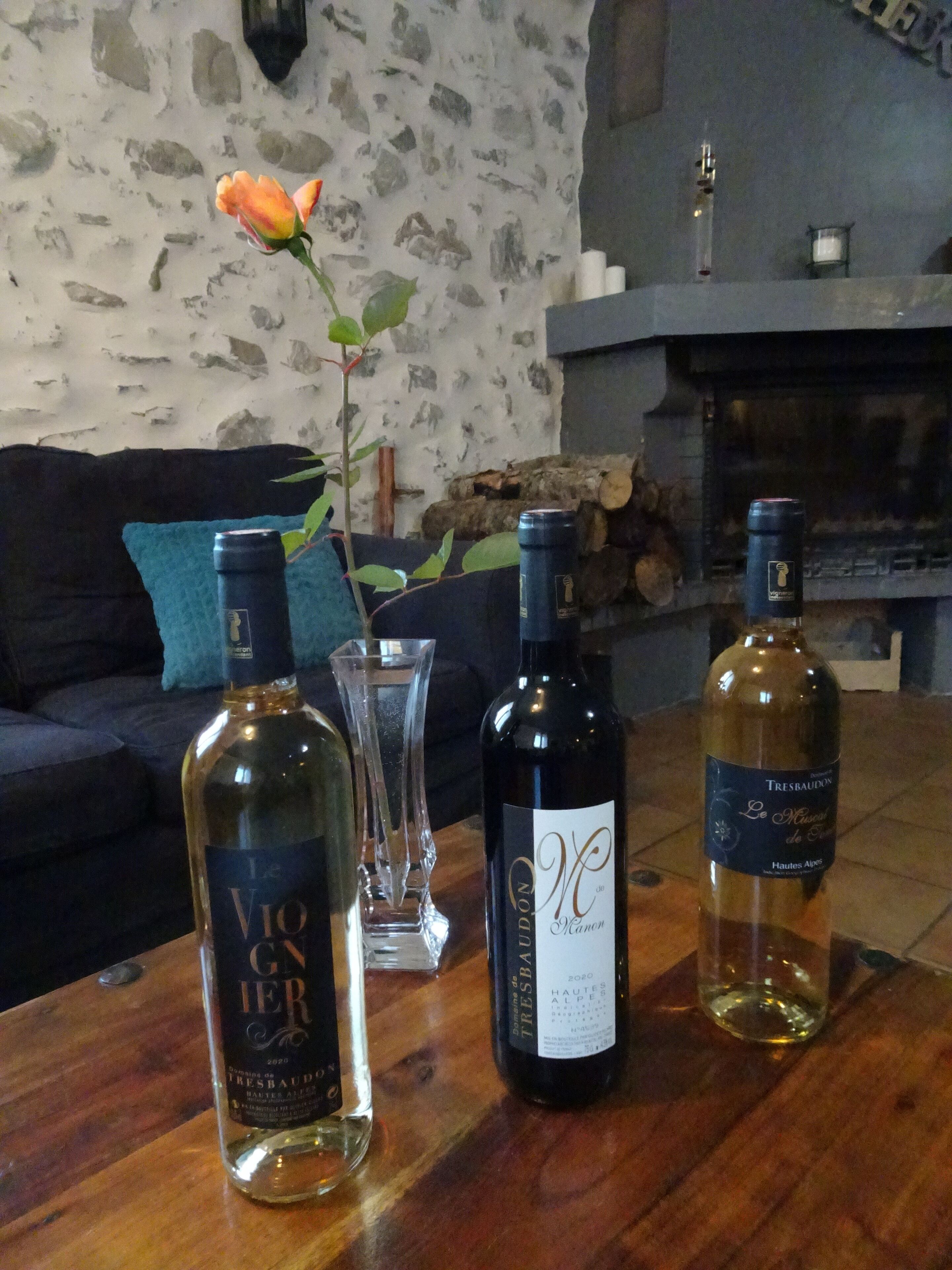 Une table en bois, avec trois bouteilles de vin, un vase contenant une seule fleur et, en arrière-plan, un mur de pierres.