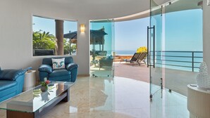 Ein modernes Wohnzimmer mit Glaswand und Meerblick, ein blaues Sofa und ein Glastisch.