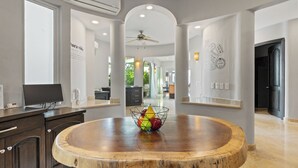 Un hall d’entrée spacieux, avec une table ronde en bois au centre, un ventilateur de plafond et, au fond, un espace cuisine moderne.