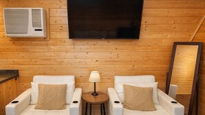 A room with two white sofas, a small round table with a lamp, a large flat-screen TV mounted on the wall, and a wooden floor. - Beautiful Log cabin overlooking a meadow (Pipe Creek)