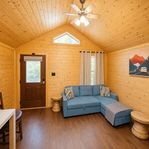 L’intérieur chaleureux d’un chalet avec un plafond en bois, un ventilateur de plafond, un canapé d’angle bleu, une table à manger et un tableau accroché au mur.