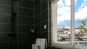 Un baño con baldosas oscuras, una ducha, una ventana con vistas a edificios y montañas, y un perchero para toallas.