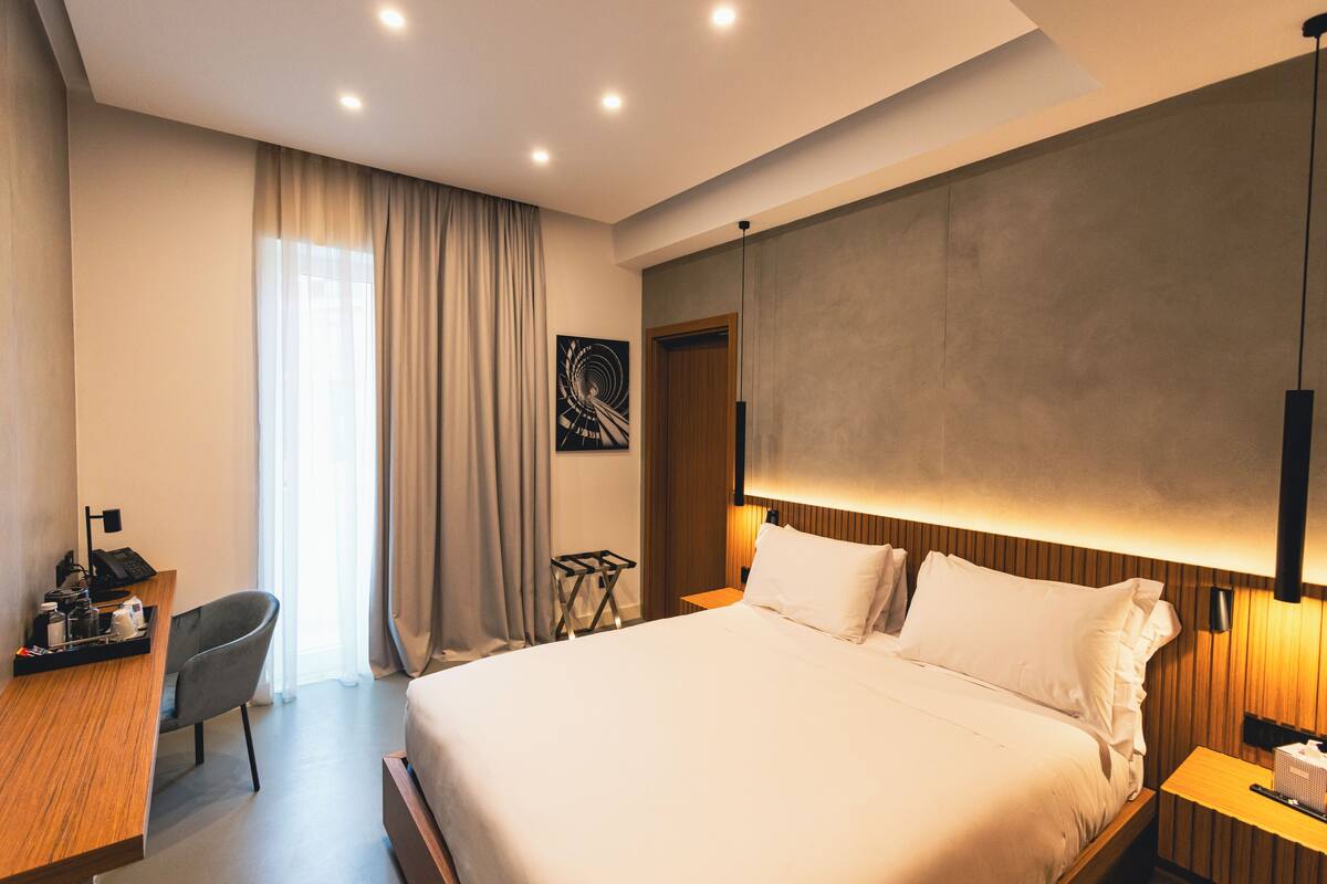 Habitació d'hotel moderna amb un llit gran, un escriptori amb una cadira, una taula petita i una finestra ampla amb cortines.