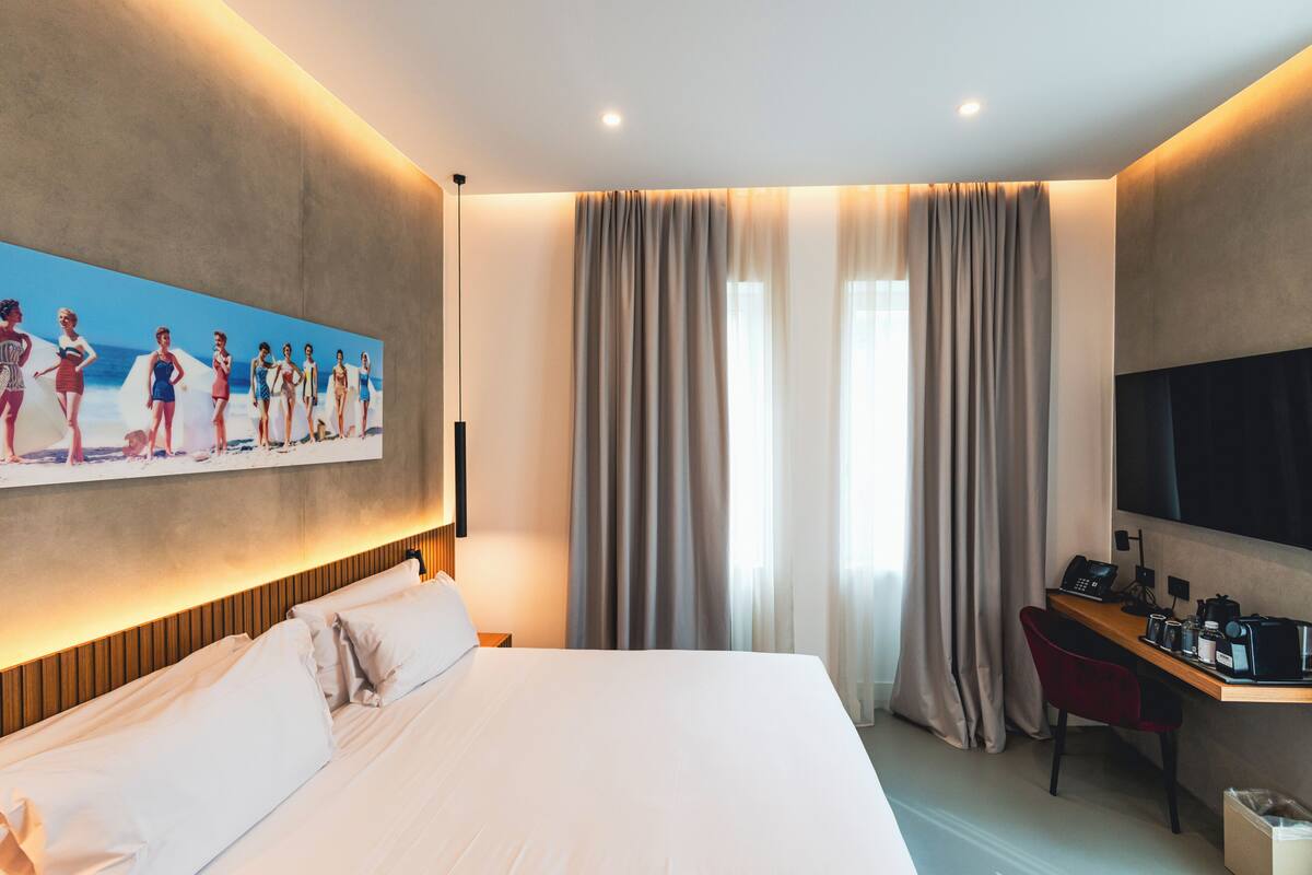 Una habitació d'hotel amb un llit gran, una taula de treball amb un ordinador, un televisor i un quadre de persones a la platja.