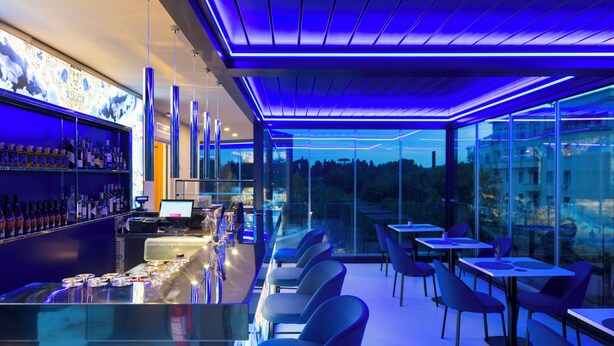 A modern bar area with a glass counter, seating, and a view of the outdoors. - J Maluni City Hotel (Rome)