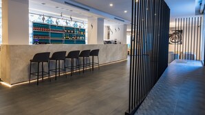 A modern bar area with a long counter, multiple bar stools, and a well-lit space with a decorative wall and ceiling lights. - J Maluni City Hotel (Rome)