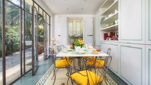A dining area with a table set for four, surrounded by chairs with yellow cushions, a kitchen area with white cabinets, and a view through large glass doors to a garden. - Villa Gaius 8+2, Emma Villas (Rosignano Marittimo)