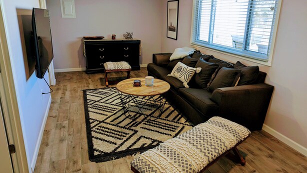 Un salon comprenant un canapé, une table basse et un pouf. Un téléviseur est fixé au mur, il y a une fenêtre avec des stores et un tapis à motifs.