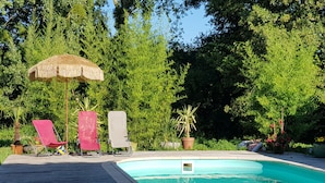 A swimming pool with a wooden deck, two red lounge chairs, a thatched umbrella, and lush greenery in the background.