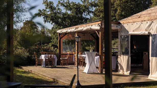 An outdoor wooden deck with tables and chairs, a pergola with hanging lights, and a covered area with a table.