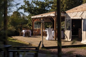 Une terrasse extérieure en bois, dotée de tables et de chaises, une pergola agrémentée de guirlandes lumineuses et un espace couvert avec une table.
