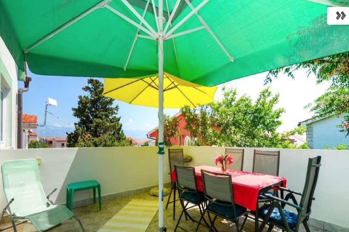 A patio with a green and yellow umbrella, a table with chairs, and a view of buildings and trees.