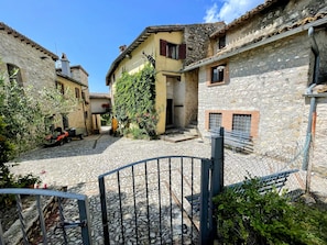 Une cour pavée de pierres, bordée de bùtiments en pierre, avec une porte en métal et un banc.