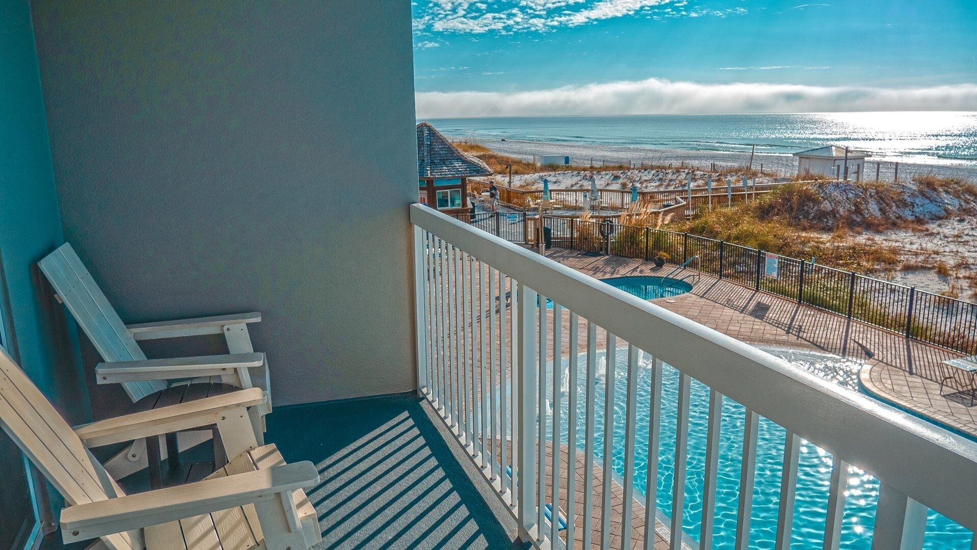 A balcony with a view of the ocean, a pool, and beach chairs.