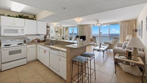 A modern kitchen with white cabinetry, a granite countertop, and a central island. There is a dining area with a sofa and a table, and a balcony with a view of the ocean. - Pelican Beach 1202 1 Bedroom Condo by Pelican Beach Management (Destin)